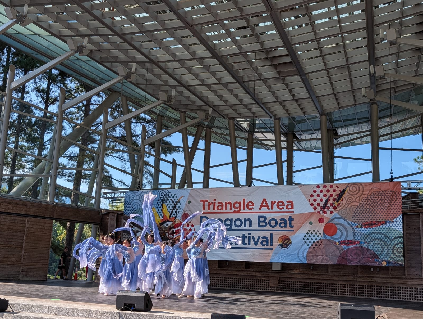 Young dancers in flowing pale-blue costumes perform a classical Chinese dance on the festival stage beneath the Triangle Area Dragon Boat Festival banner