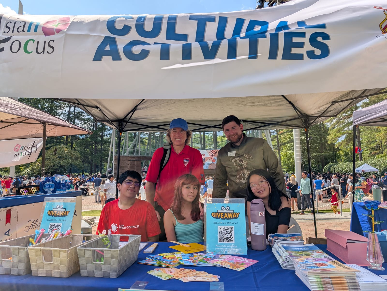 Three teenagers seated at the Asian Focus Cultural Activities survey table with giveaway materials, two adults standing behind them