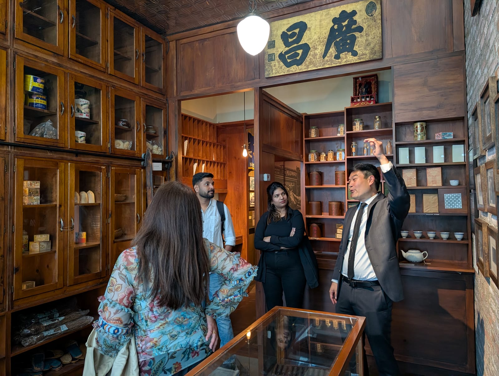 Museum president Michael Lee showing a recreated century-old Chinatown general store, its wooden cabinets stocked with jars and goods beneath a gilded Chinese shop sign