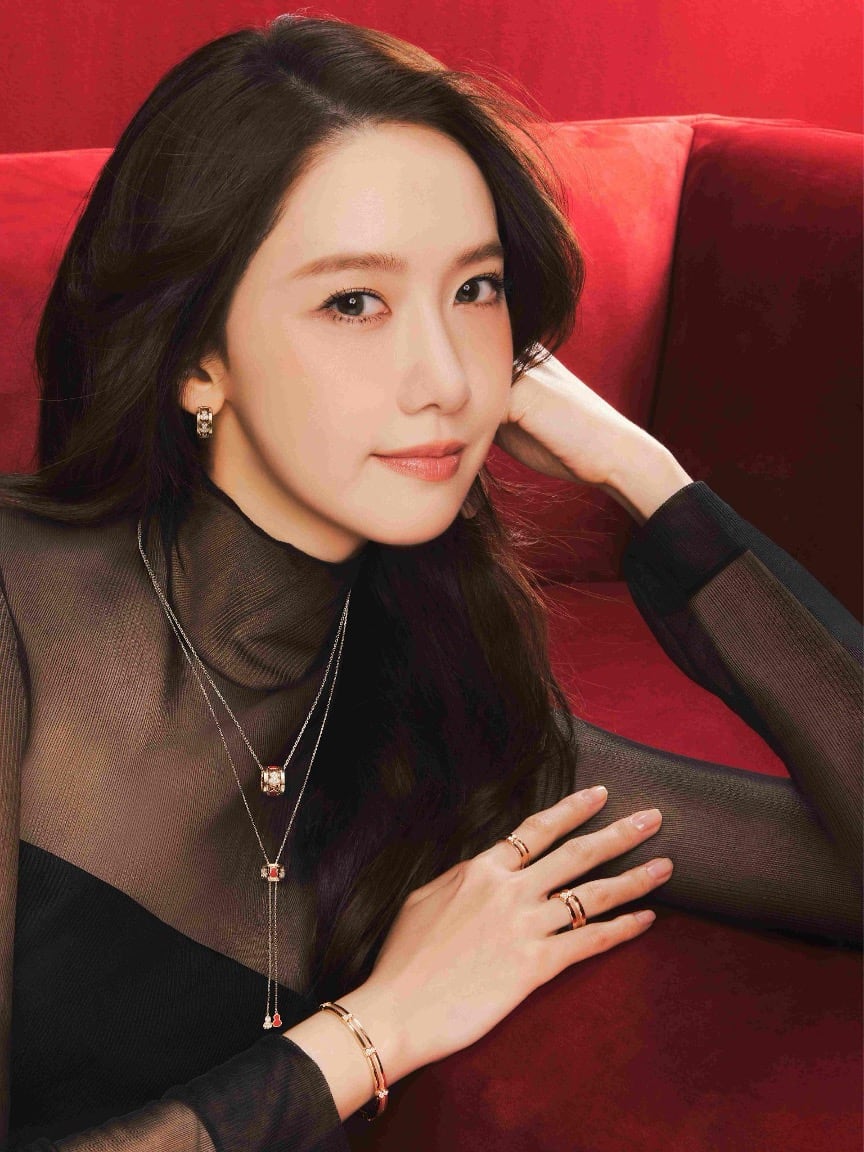 A model reclining on a red couch wearing Qeelin fine jewelry — a layered necklace, rings, and bracelet